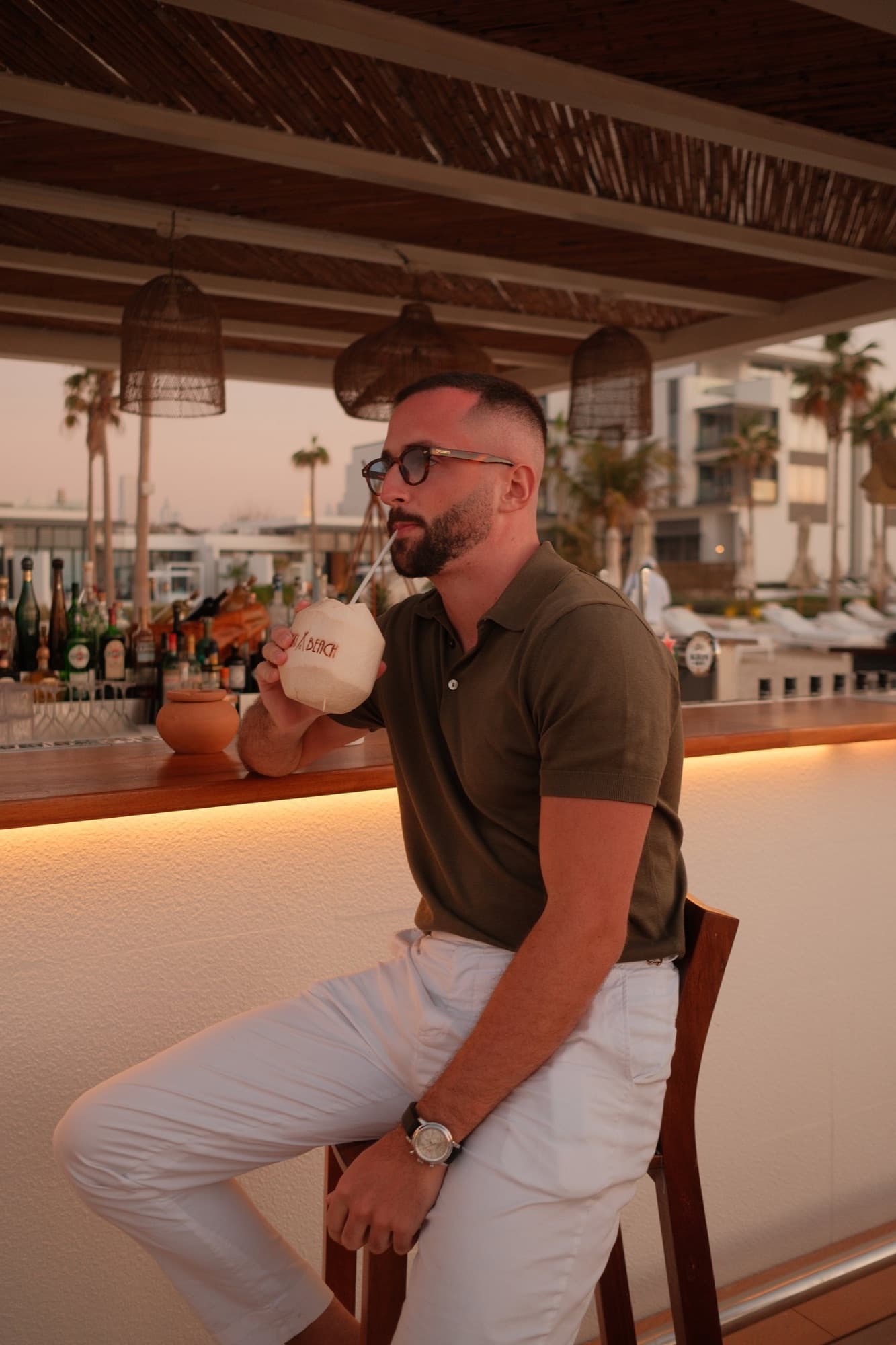 Ein Mann mit Sonnenbrille und grünem Polohemd trinkt aus einer Kokosnuss an einer Bar im Freien.