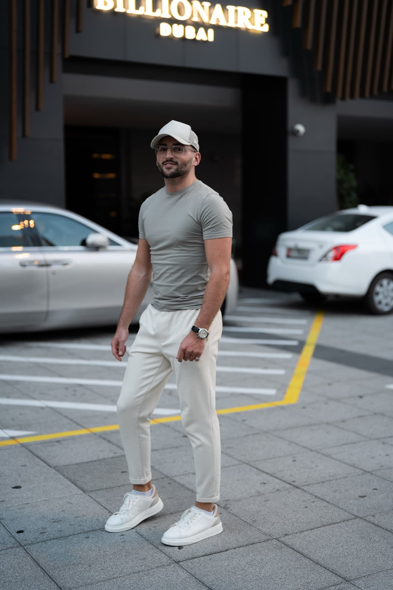 Ein Mann in beiger Kappe, T-Shirt, heller Hose und weißen Turnschuhen steht vor einem Gebäude mit der Aufschrift „BILLIONAIRE DUBAI“.