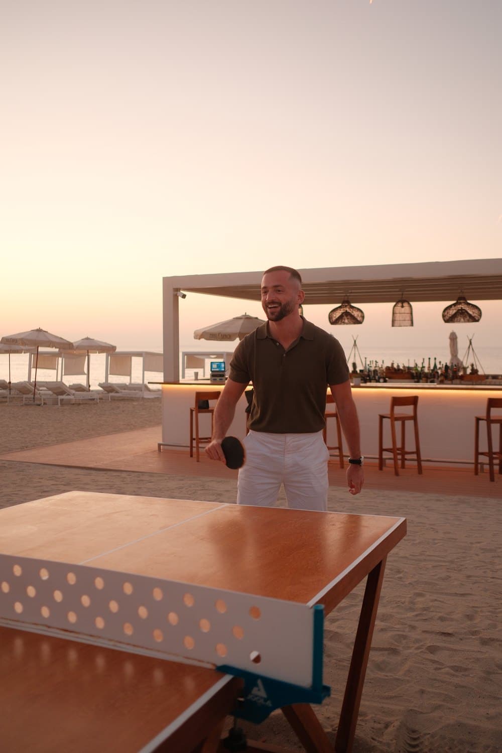 Ein lächelnder Mann spielt Tischtennis am Strand bei Sonnenuntergang vor einer Bar.