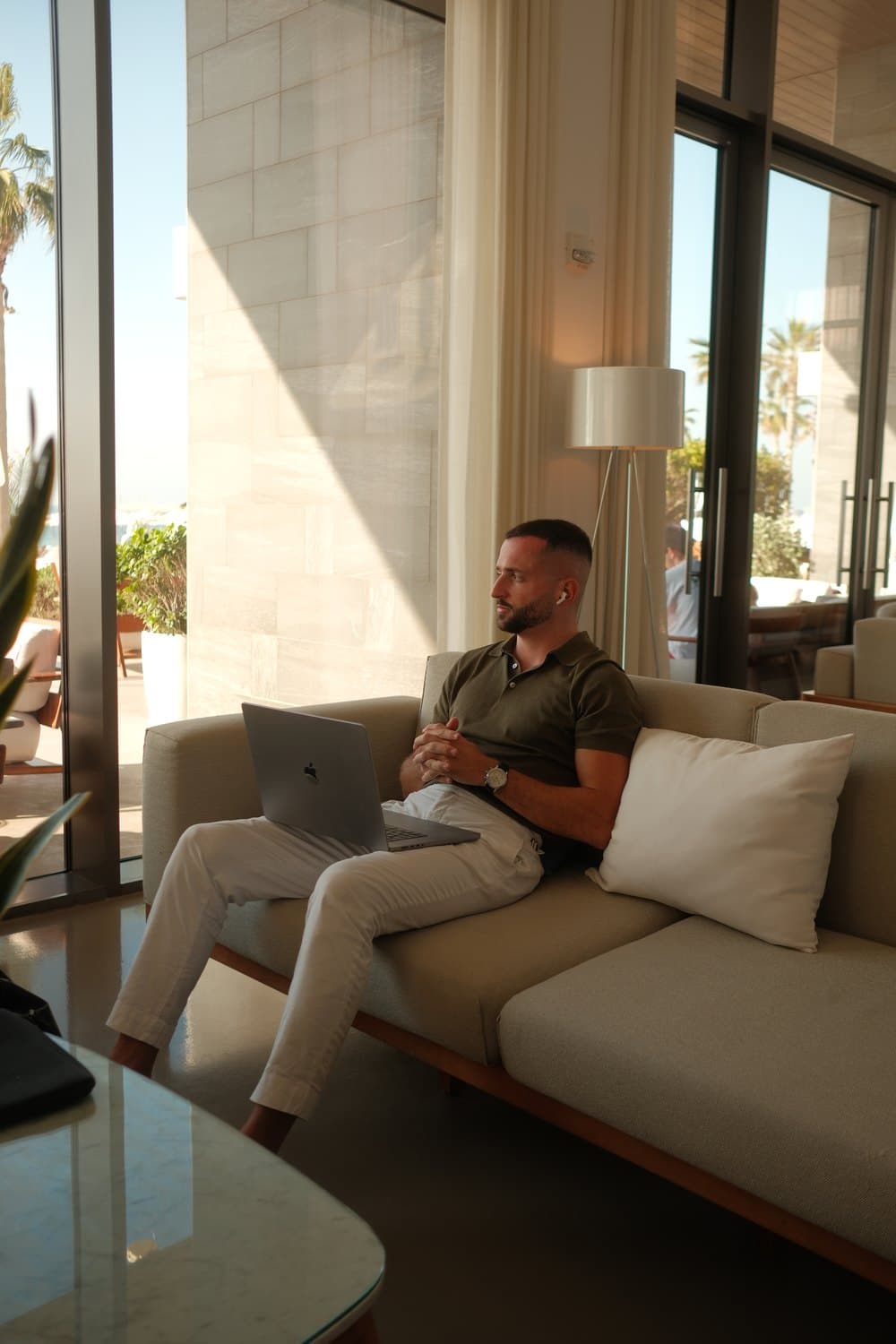 Mann mit Laptop auf beigefarbenem Sofa in sonnigem Raum mit großen Fenstern.