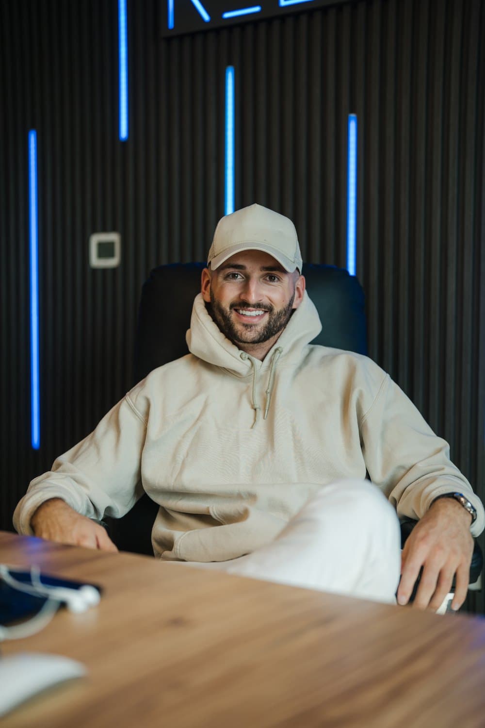 Ein lächelnder Mann mit beigem Hoodie und Baseballkappe sitzt an einem Holztisch vor einer dunklen Wand mit blauen Lichtstreifen.