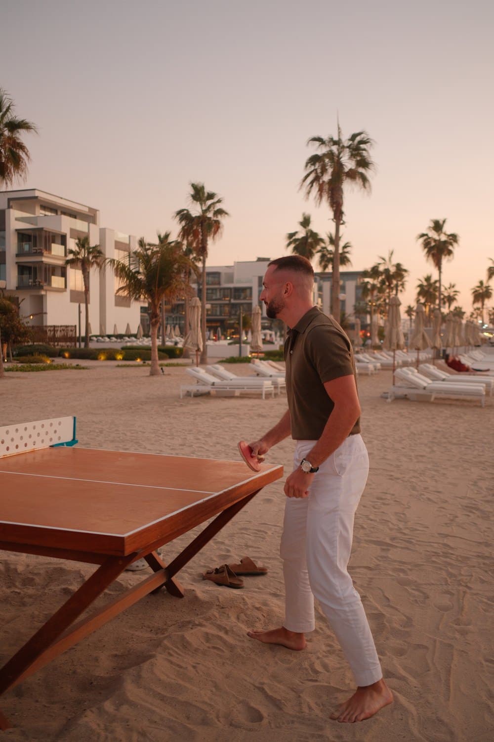 Ein Mann spielt barfuß Tischtennis am Strand bei Sonnenuntergang, mit Palmen und Gebäuden im Hintergrund.